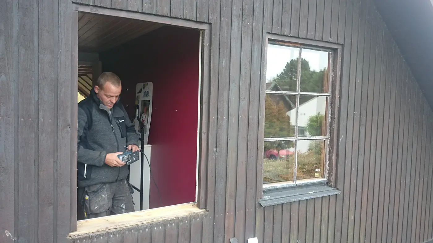 En man står vid ett öppet fönster i en träbyggnad och tittar på en apparat som han håller i händerna.