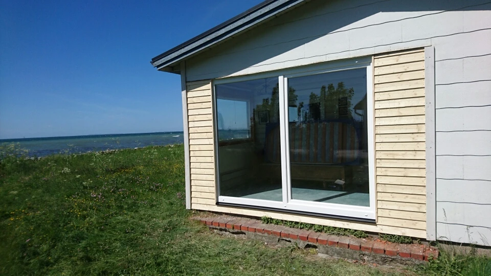 Ett litet vitt hus med ett stort fönster med utsikt över havet och det gröna landskapet.